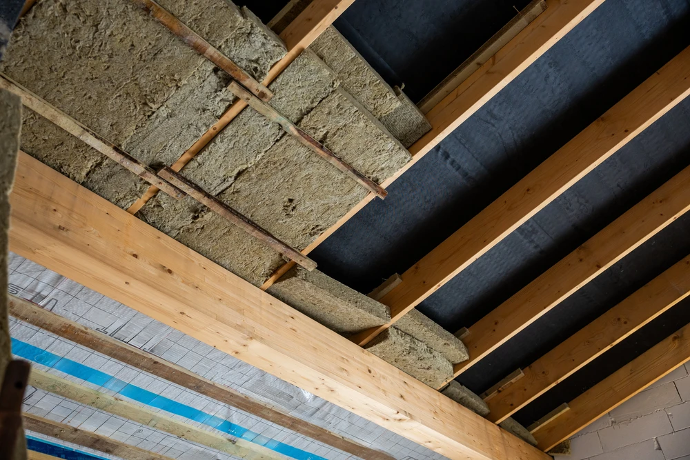 Les poutres en bois apparentes et le matériau isolant sont visibles dans un bâtiment en construction, avec un pare-vapeur noir sur la section du plafond