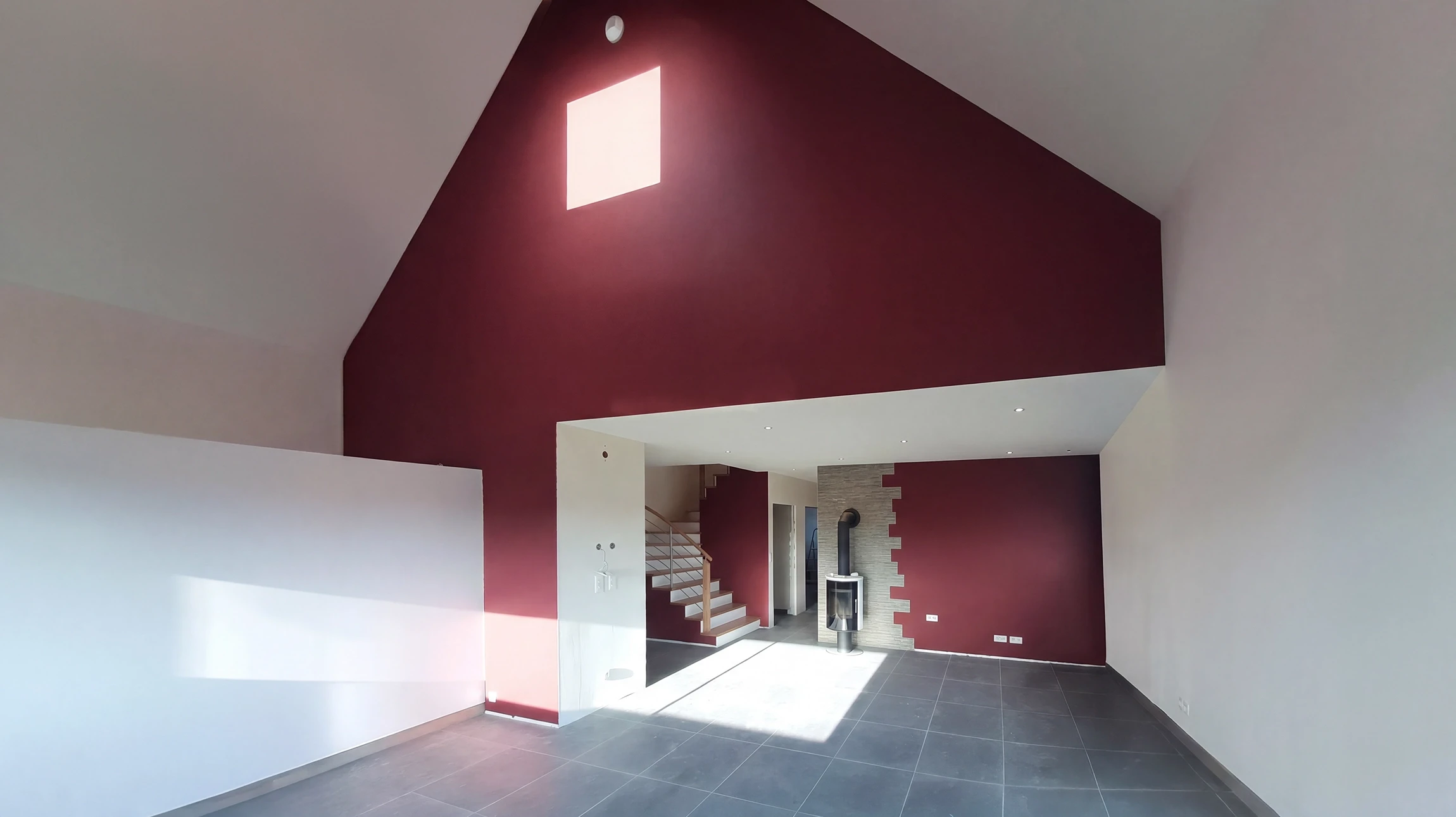 Photographie d'un artisan en tenue de travail appliquant du papier peint ou un revêtement mural dans un intérieur résidentiel. L'artisan doit être saisi en train de lisser ou poser le revêtement sur un mur, montrant le professionnalisme et la technique. La scène se déroule dans une pièce en cours de rénovation, avec des outils visibles (lisseur, couteau à enduire, etc.). L'éclairage naturel doit mettre en valeur la qualité du travail effectué.